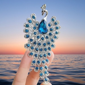 Elegant peacock brooch with blue crystals and silver finish held against a sunset ocean backdrop
