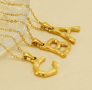 Close-up of gold stainless steel initial pendants A, B, and C on beige fabric background