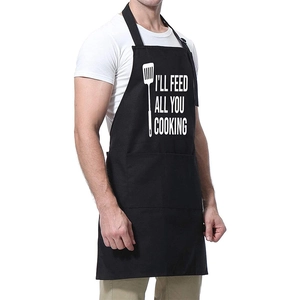 Detail der schwarzen Schürze mit weißem Aufdruck I'LL FEED ALL YOU COOKING und Pfannenwender