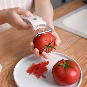 White kitchen vegetable peeler with metal blade peeling a fresh tomato on a wooden table