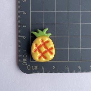 Detail shot of a yellow resin pineapple charm showing precise 2.5cm size on a black scale