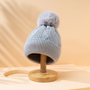 Light grey knitted beanie with fluffy pom pom on wooden stand under soft tree shadows