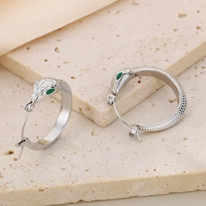 Silver snake hoop earrings with green gemstone eyes and scales on beige stone background