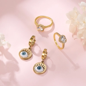 Golden Evil Eye earring set and zircon rings on a soft pink background with flowers