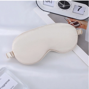 Beige faux silk sleep mask displayed with an alarm clock and magazine on soft bedding