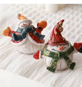 Close-up of two miniature resin snowmen with festive hats and scarves on snowy fabric