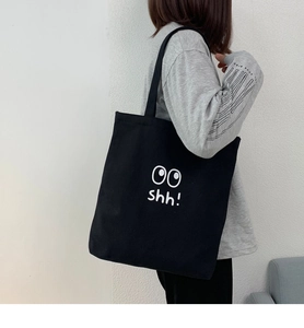 Detalle de un bolso tote de lona negra con texto shh! blanco y gráfico de unos ojos