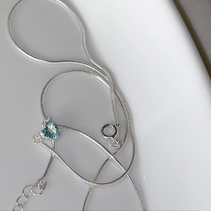 Detail of silver snake chain necklace featuring a light blue heart-shaped gemstone pendant