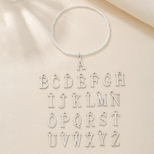 Set of 26 silver initial pendants and white seed bead anklet on a neutral textured background