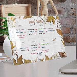Close-up of a clear acrylic weekly planner board with green leaf design on a wooden desk