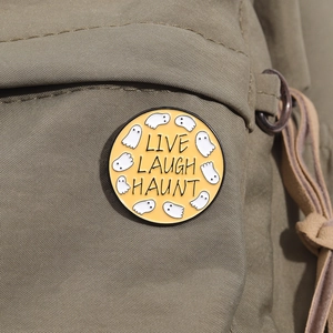 Detail showing round enamel ghost brooch on beige canvas backpack with text LIVE LAUGH HAUNT