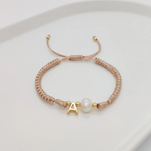 Close-up of handmade woven bracelet with white pearl and gold letter A charm on tray