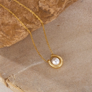 Close-up of a gold U-shaped pendant necklace featuring a white pearl on a stone surface