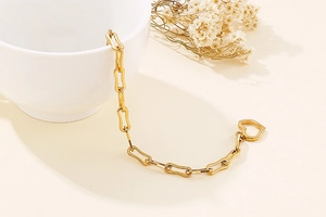 Close-up of golden paperclip chain bracelet with heart toggle clasp on a beige background