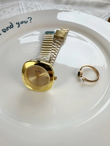 Detail of a square gold watch with mesh strap and a matching black clover ring on a plate