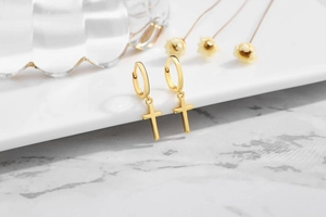 Macro detail of gold cross charm drop earrings on a minimalist white pedestal display
