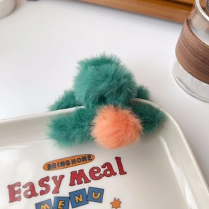 Detailed close-up of a green plush hair claw with orange pom-pom on a ceramic plate