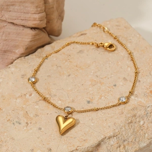 Detail of a gold heart charm anklet with sparkling zircon stones on a beige stone surface