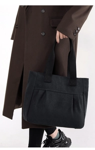 Detail of black canvas tote bag with pleated front pocket held by model in trench coat