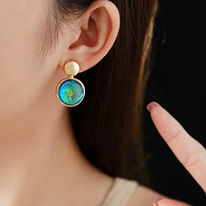 Close-up of golden circular earrings with iridescent blue resin stones worn by a model