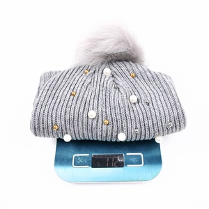 Berretto in maglia grigio con perle e pompon su bilancia che indica 112g di peso