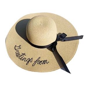 Beige straw sun hat with Greetings from embroidery and black ribbon bow on white background