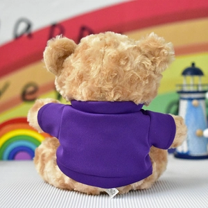 Back view of a light brown plush teddy bear wearing a blank purple t-shirt.