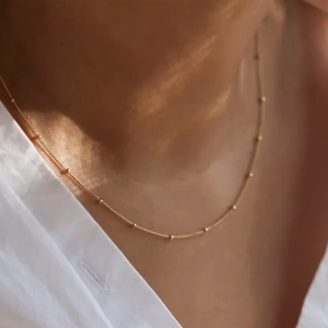 Close-up of a minimalist gold beaded chain necklace worn by a model with a soft focus background