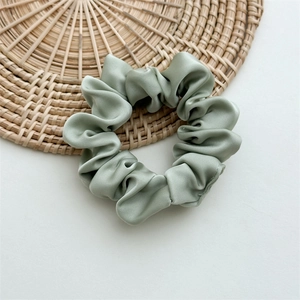 Close-up of a sage green satin scrunchie featuring elegant folds on a woven mat