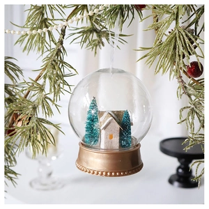 Close-up of a Christmas snow globe ornament with a white cabin and golden base on a pine branch
