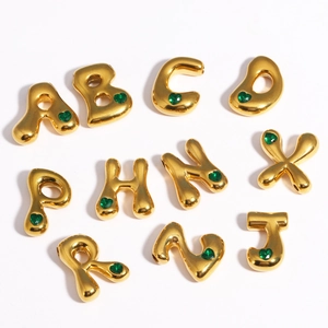 Détail de breloques lettres dorées avec gemmes cœur vert sur fond blanc épuré