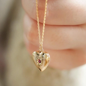 Detailed view of a gold heart locket necklace with a red birthstone held by a hand
