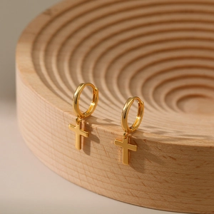 Detail of gold-plated cross dangle hoop earrings hanging on a minimalist wooden stand