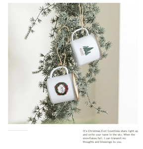Close-up of mini white enamel Christmas cup ornaments with tree and wreath on pine branch