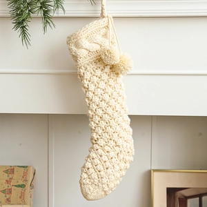 Cream knit Christmas stocking with pom-poms and tassels hanging by a cozy fireplace
