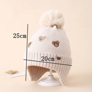 Detail of beige knit winter hat showing bear embroidery and large faux fur pom-pom