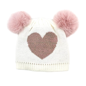 Detail of cream knit baby hat featuring pink glitter heart and soft dual pom-poms