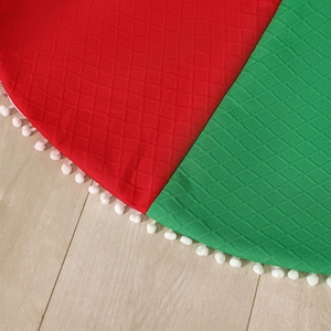 Red and green quilted Christmas tree skirt with white pom-pom trim on light wood floor