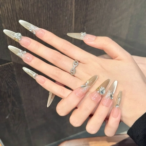 Dettaglio di unghie stiletto con finitura cat-eye champagne, perle e fiocchi in 3D