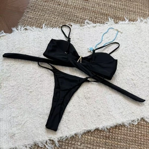 Flat lay of black bikini set featuring a gold V-shaped metal link on white fringe carpet