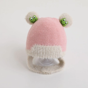 Detailed close-up of pink knitted baby hat with white faux fur balls and green frog patch