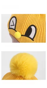 Detail of yellow knit winter hat featuring cartoon eye embroidery and soft faux fur pompom