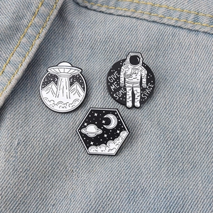 Space-themed black and white enamel pins set on denim featuring astronaut and UFO designs