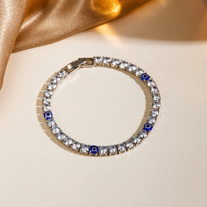 Detail of tennis bracelet featuring white zircons and blue evil eye charms on beige background