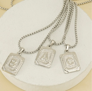 Three silver square initial necklaces with A, B, and C pendants on a white stone stand