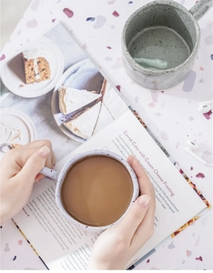 Close-up of hands holding a grey speckled ceramic mug with coffee over an open recipe book