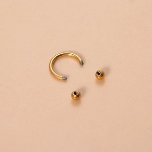 Golden titanium horseshoe circular barbell piercing with screw balls on beige background