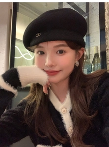 Detail of a black wool beret featuring a metallic M emblem worn with a knit sweater