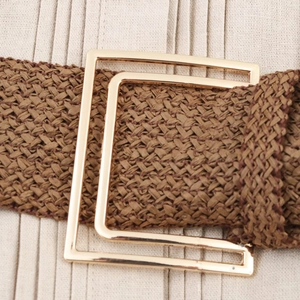 Detail of a brown woven belt featuring a polished gold square metal buckle on beige fabric