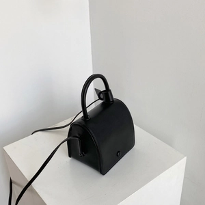 Minimalist black leather crossbody bag with top handle on white pedestal display